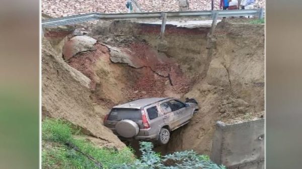 Car stranded in a collapsed hillside under a highway barrier, buried up to the wheels by mud and soil with bystanders nearby (informative image).