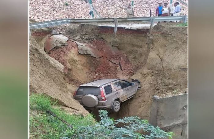 Car stranded in a collapsed hillside under a highway barrier, buried up to the wheels by mud and soil with bystanders nearby (informative image).