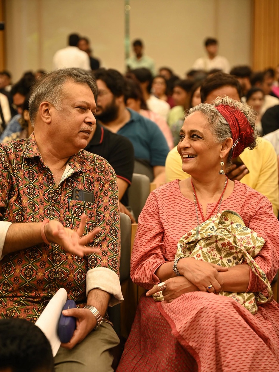 Two adults in a crowded hall share a smile while seated together, chatting at an event.