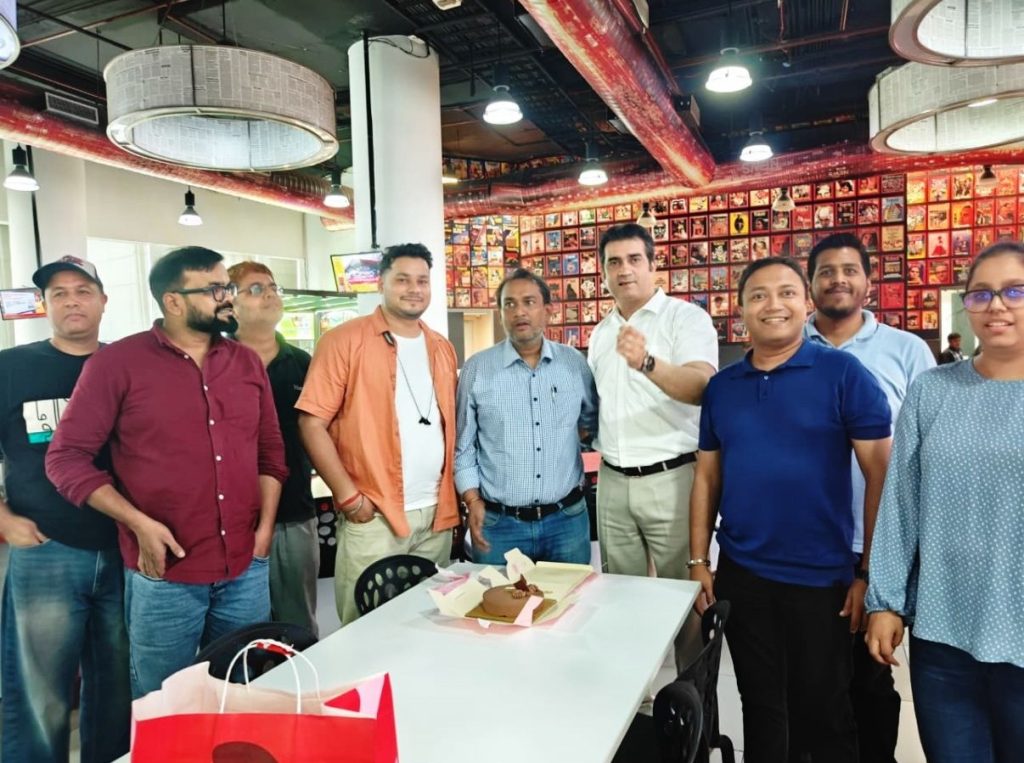 prashant | No. 1 Indian Media News Portal A group of people standing around a table with a small cake and gift bags in a bright, modern office or cafe, with a wall of photos in the background.
