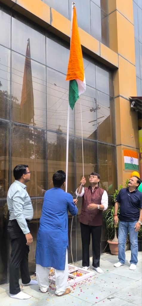 Group of men outdoors hoisting the Indian national flag on a tall pole in front of a glass-fronted building during a ceremony.