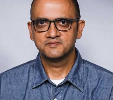 Portrait of a man with short hair and glasses, wearing a blue denim shirt, looking at the camera against a light gray background.