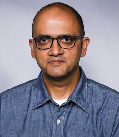 Portrait of a man with short hair and glasses, wearing a blue denim shirt, looking at the camera against a light gray background.