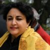 Woman with short curly black hair wearing a yellow patterned shawl, standing outdoors with a thoughtful expression.