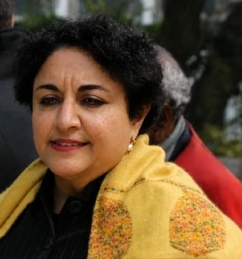 Woman with short curly black hair wearing a yellow patterned shawl, standing outdoors with a thoughtful expression.