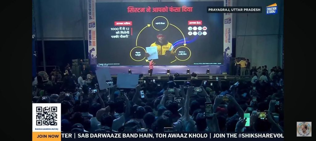 Speaker on a stage presenting to a packed indoor audience, with a large slide screen behind and people filming with phones; QR code at left bottom and ticker text at bottom edge.