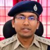 Police officer in a khaki uniform and blue beret speaking at the camera, red curtains behind him.