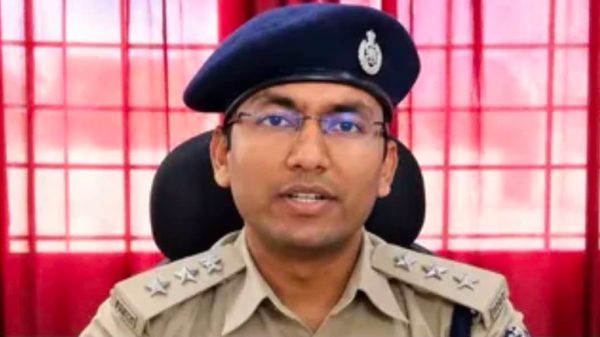 Police officer in a khaki uniform and blue beret speaking at the camera, red curtains behind him.