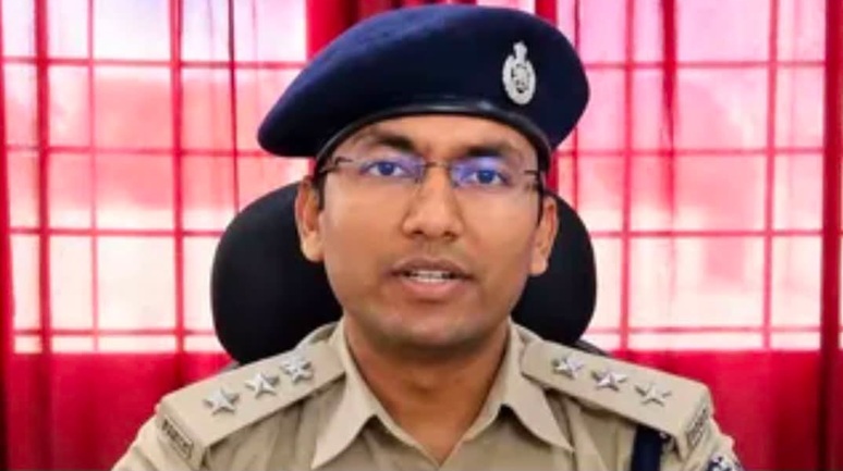 Police officer in a khaki uniform and blue beret speaking at the camera, red curtains behind him.