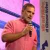Man in a pink shirt speaks into a handheld microphone on a stage with a pink backdrop.