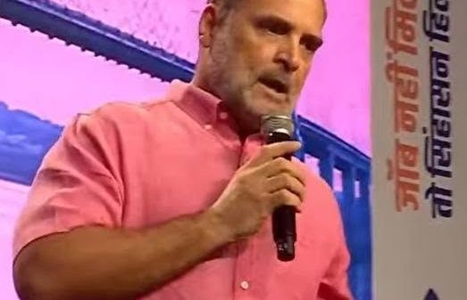Man in a pink shirt speaks into a handheld microphone on a stage with a pink backdrop.