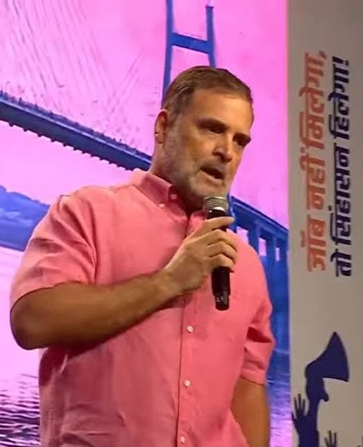 Man in a pink shirt speaks into a handheld microphone on a stage with a pink backdrop.
