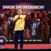 Speaker in a yellow shirt on stage addressing a large crowd with the banner 'SMASH THE PATRIARCHY' behind him on a red-carpeted stage.