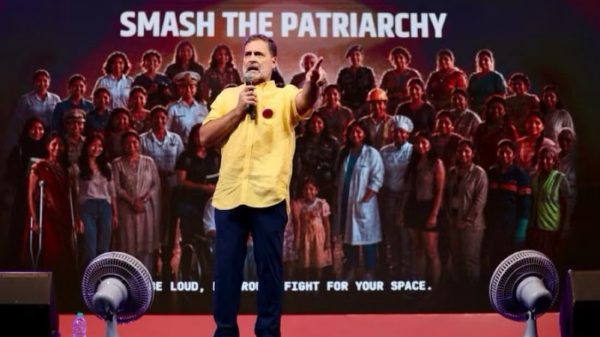 Speaker in a yellow shirt on stage addressing a large crowd with the banner 'SMASH THE PATRIARCHY' behind him on a red-carpeted stage.