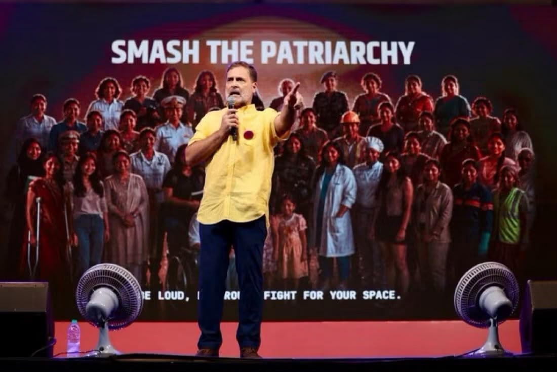 Speaker in a yellow shirt on stage addressing a large crowd with the banner 'SMASH THE PATRIARCHY' behind him on a red-carpeted stage.