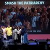 Group of people on a stage posing for a photo at an event, with a large banner reading 'SMASH THE PATRIARCHY' in the background and a TV-style 'LIVE' graphic on the right.