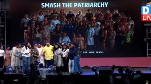 Group of people on a stage posing for a photo at an event, with a large banner reading 'SMASH THE PATRIARCHY' in the background and a TV-style 'LIVE' graphic on the right.