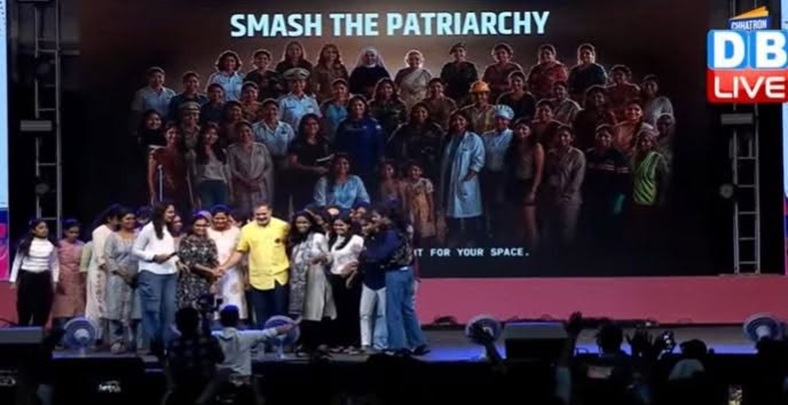 Group of people on a stage posing for a photo at an event, with a large banner reading 'SMASH THE PATRIARCHY' in the background and a TV-style 'LIVE' graphic on the right.