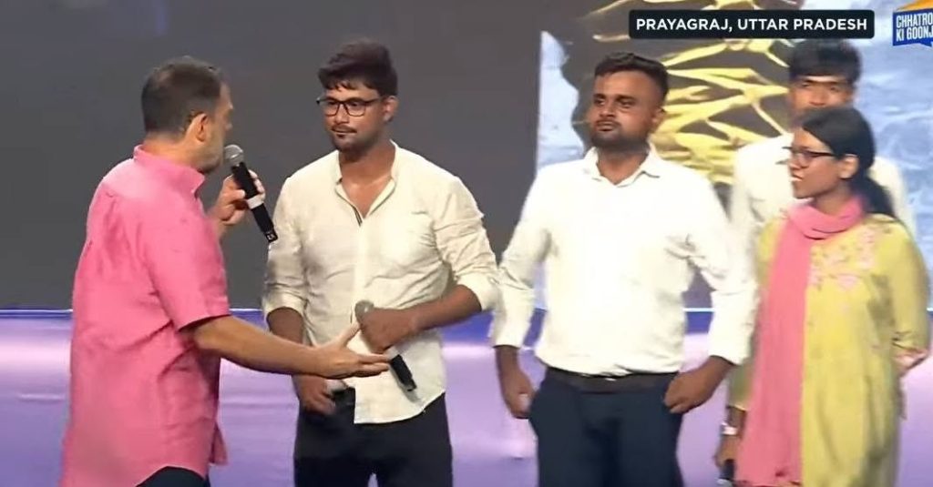 Man in pink shirt hands a microphone to a man in glasses on a stage, with two others observing. Location: Prayagraj, Uttar Pradesh.