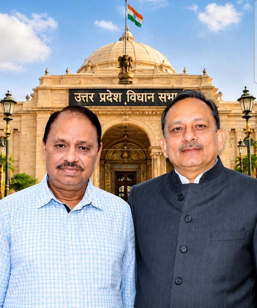 Two men pose in front of the Uttar Pradesh Legislative Assembly building, with a domed facade, ornate arches, and the Indian flag overhead.