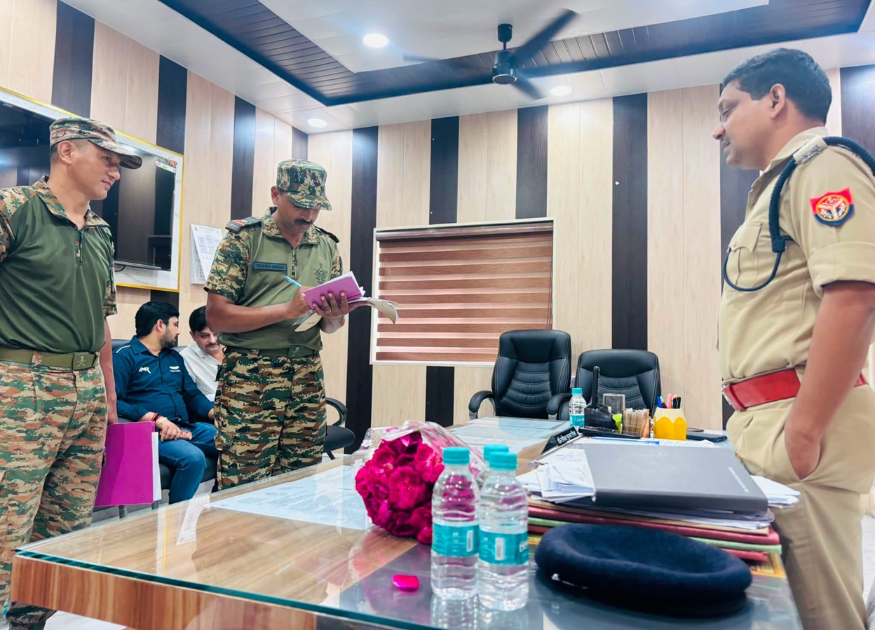 Three uniformed officers in a conference room, two in camouflage and one in a tan uniform, standing around a glass table with papers and bottles as colleagues sit in the background.