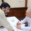 Author in a white shirt signs a book at a table, leaning toward a man presenting a copy.