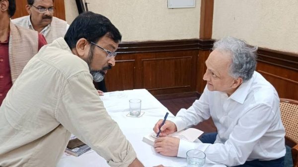 Author in a white shirt signs a book at a table, leaning toward a man presenting a copy.