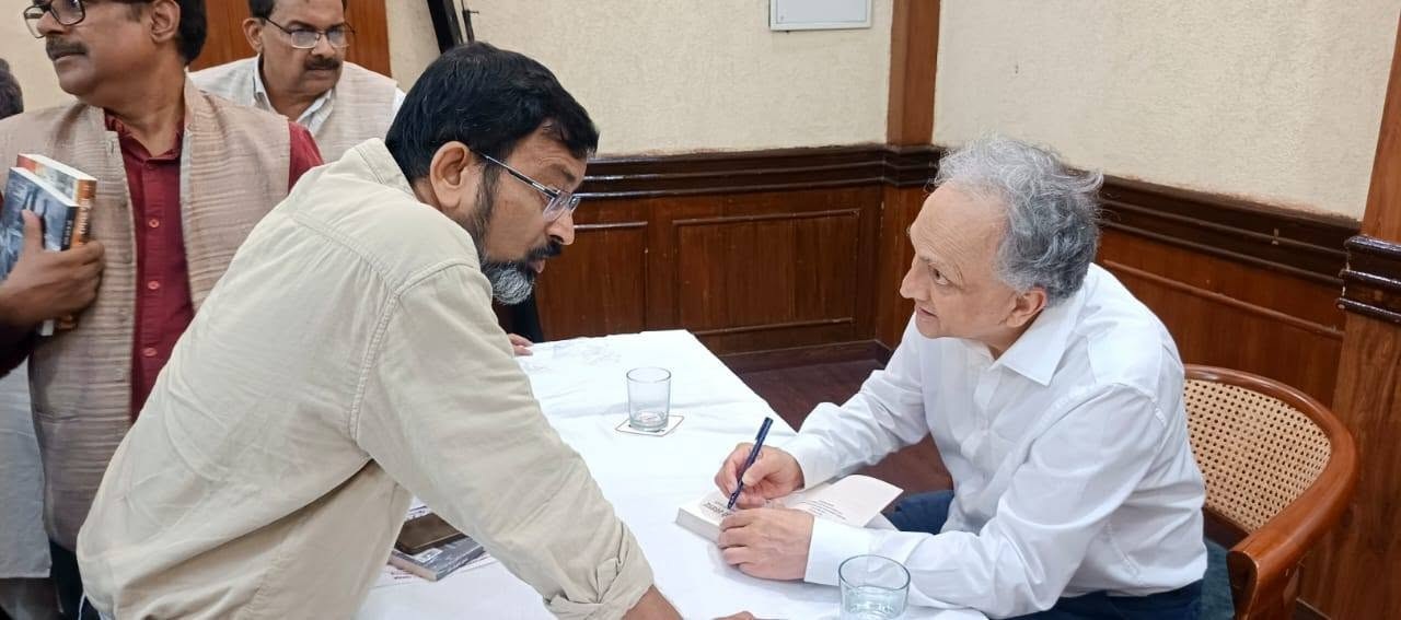 Author in a white shirt signs a book at a table, leaning toward a man presenting a copy.