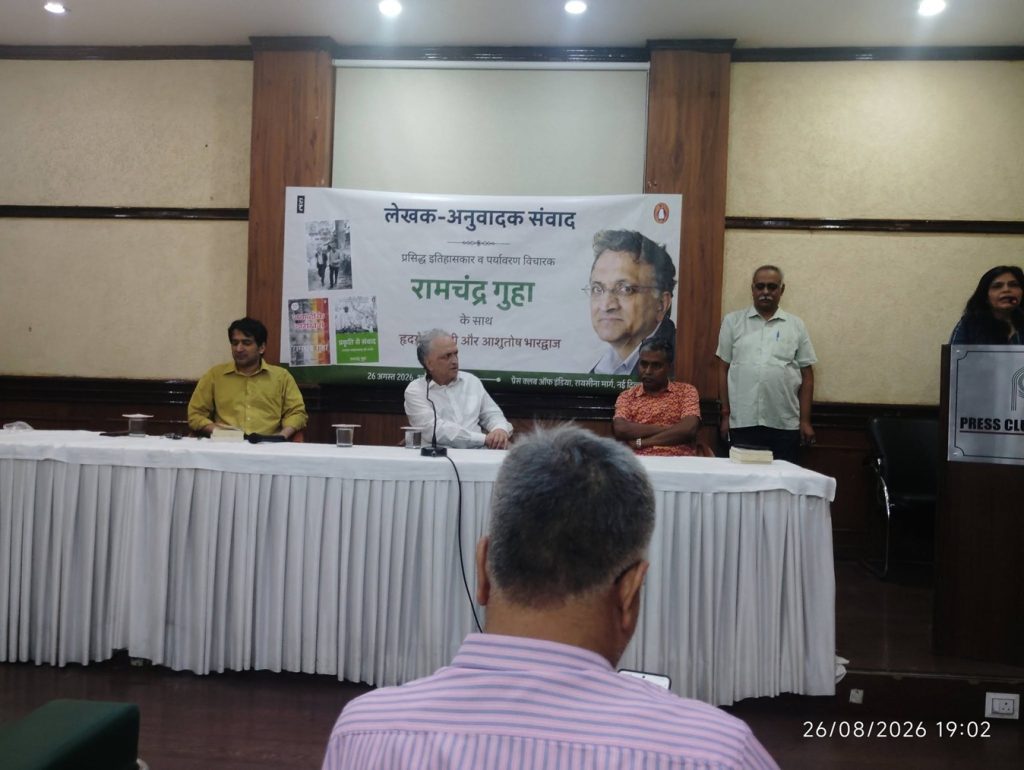 ramchandra guha_1 | No. 1 Indian Media News Portal Panel discussion at a literary event; four men seated behind a long table on a stage with a banner featuring Ramchandra Guha in hardcover. Behind them is a large poster and a portrait image.
