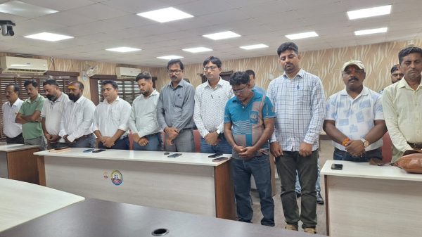 Group of men standing in a row behind conference desks in a meeting room, heads bowed.