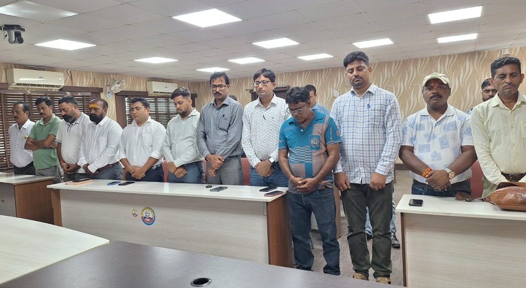 Group of men standing in a row behind conference desks in a meeting room, heads bowed.