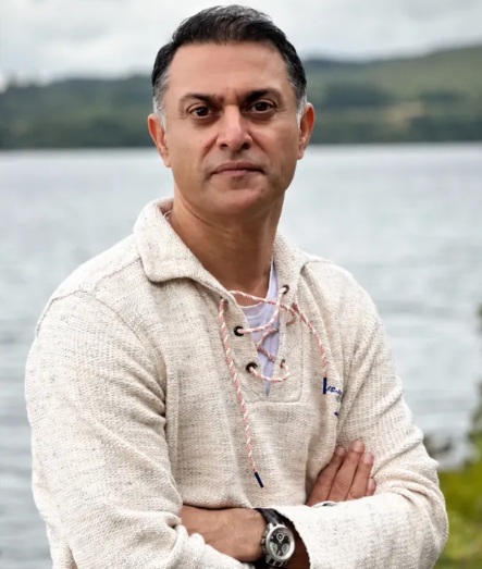 rohit gopakumar | No. 1 Indian Media News Portal Man with short dark hair wearing a cream knit sweater, standing with arms crossed by a lake with hills in the background.