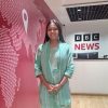 Smiling woman in a mint green traditional outfit stands in a BBC News lobby with a pink wall full of text and a reception desk nearby.