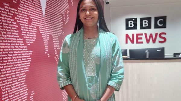 Smiling woman in a mint green traditional outfit stands in a BBC News lobby with a pink wall full of text and a reception desk nearby.