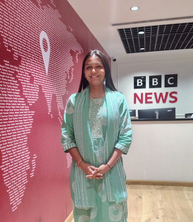 Smiling woman in a mint green traditional outfit stands in a BBC News lobby with a pink wall full of text and a reception desk nearby.