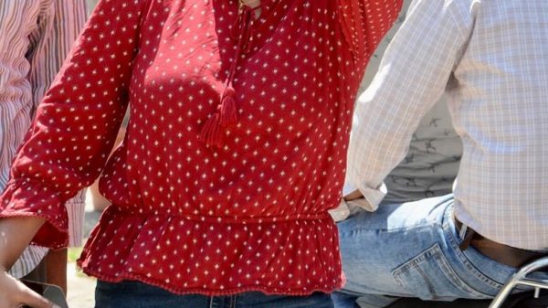 Woman in a red polka-dotted blouse and sunglasses smiles and flashes a peace sign toward the camera outdoors.