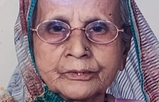 Portrait of an elderly woman wearing a multicolor headscarf and round glasses, looking at the camera.