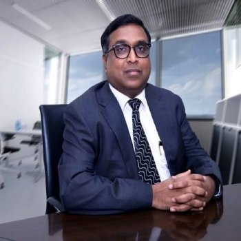 Professional portrait of a man in a navy suit and glasses, seated at a conference table in a modern office.