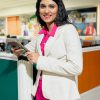 Businesswoman in a white blazer and hot pink outfit stands beside a cubicle, holding a smartphone and smiling in the office.