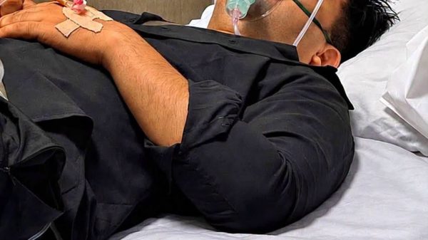 Man lying in hospital bed with an oxygen mask and IV tubing, resting with eyes closed.