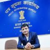 Man in a blue suit sits at a desk in front of a blue Sign: Press Information Bureau (Government of India) with the emblem above him.