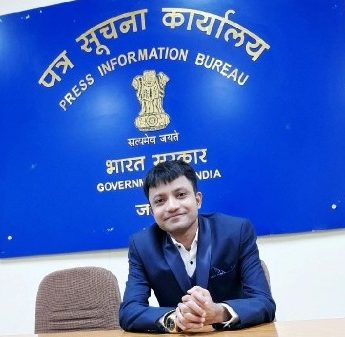 Man in a blue suit sits at a desk in front of a blue Sign: Press Information Bureau (Government of India) with the emblem above him.