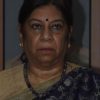 Portrait of a middle-aged Indian woman wearing a purple patterned sari with a gold border, a blue bindi, and a beaded necklace, looking straight ahead with a neutral expression.