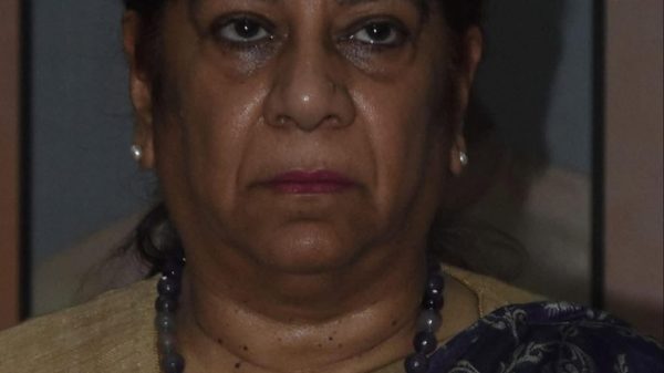 Portrait of a middle-aged Indian woman wearing a purple patterned sari with a gold border, a blue bindi, and a beaded necklace, looking straight ahead with a neutral expression.