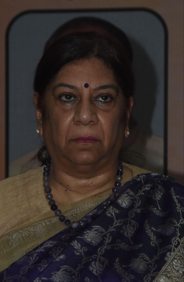 Portrait of a middle-aged Indian woman wearing a purple patterned sari with a gold border, a blue bindi, and a beaded necklace, looking straight ahead with a neutral expression.