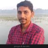 Portrait of a man with a red tilak on his forehead, wearing a red plaid shirt, standing beside a shallow water body with reeds in the background.