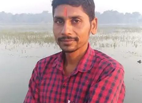 Portrait of a man with a red tilak on his forehead, wearing a red plaid shirt, standing beside a shallow water body with reeds in the background.