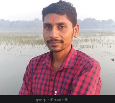 Portrait of a man with a red tilak on his forehead, wearing a red plaid shirt, standing beside a shallow water body with reeds in the background.