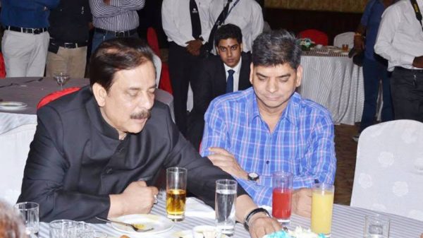 Two men sit at a banquet table, one in a dark suit and the other in a blue plaid shirt, with drinks and plates in front while people stand in the background.