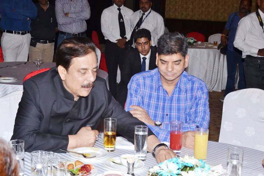 Two men sit at a banquet table, one in a dark suit and the other in a blue plaid shirt, with drinks and plates in front while people stand in the background.
